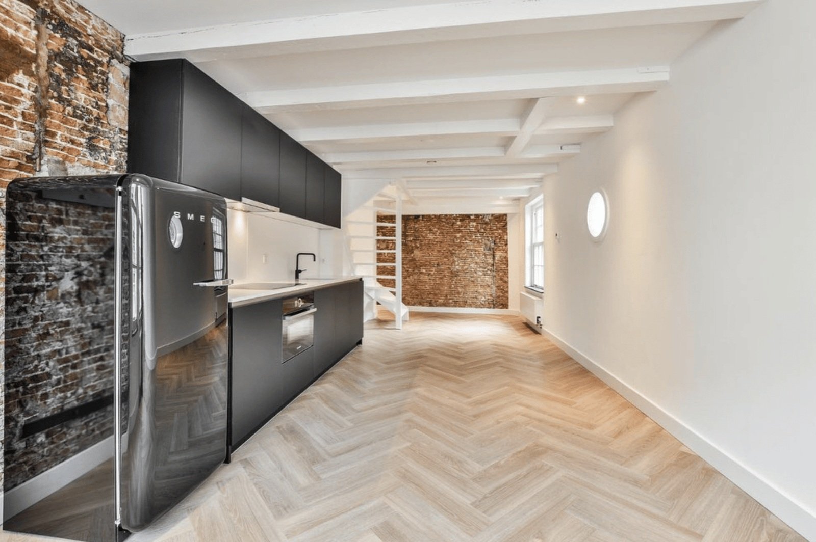 Cuisine ouverte sur un mur de brique apparente, poutres peintes et parquet en point de Hongrie