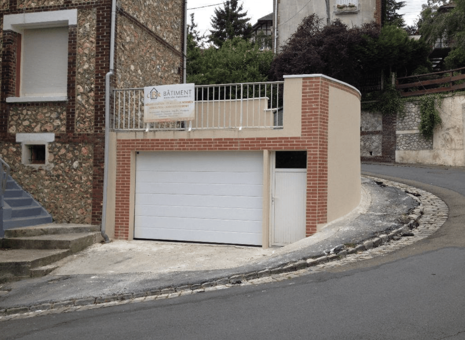 Garage et terrasse maçonnés en brique et enduit sur une maison en silex, panneau de chantier CBR en place